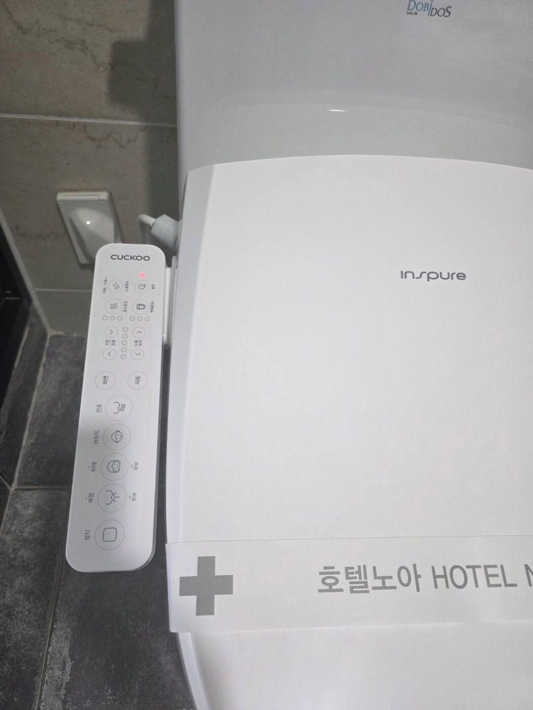 A white hotel room with a CUCKOO remote control and an INSPURE brand item, possibly a coffee maker or similar appliance.