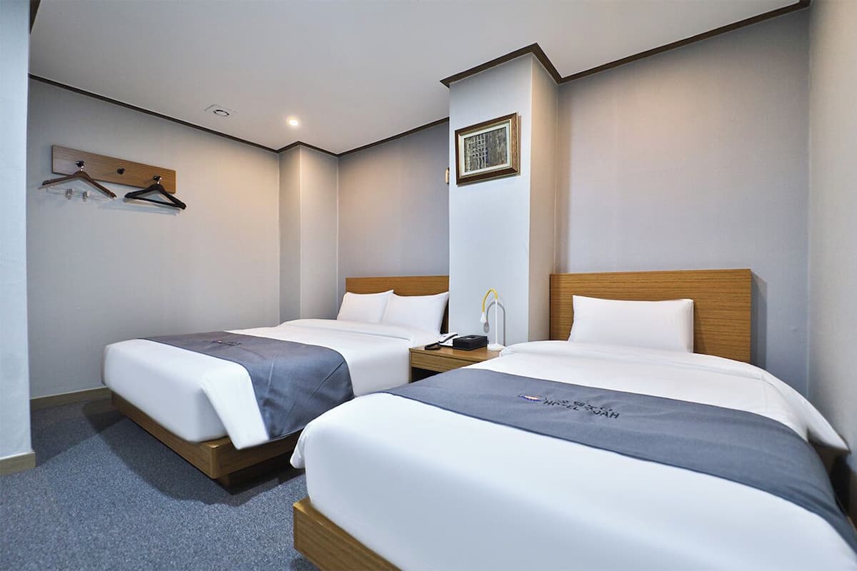 Habitación de hotel con dos camas, cabecera de madera, mesita de noche con lámpara y perchero con colgadores.