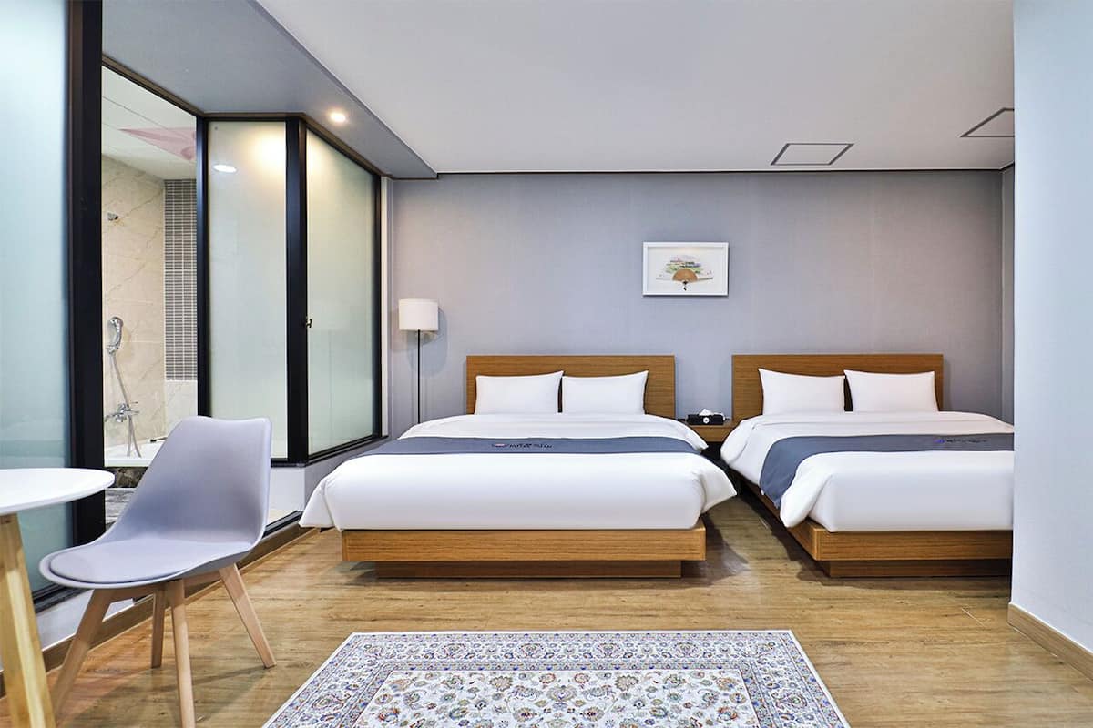 Habitación de hotel con dos camas, una silla, una mesita y un ventanal grande.