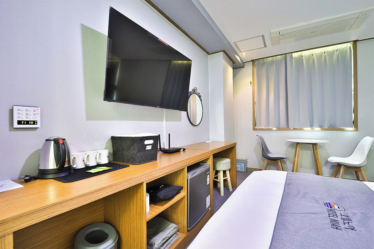 A hotel room with a bed, a desk with a kettle, cups, a mirror, a television, a small table, and a chair.