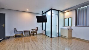 Ein modernes Zimmer mit Holzboden, einem an der Wand montierten Flachbildfernseher, einem kleinen Tisch mit Stühlen, einer Glastrennwand und einem Fenster mit Vorhängen.