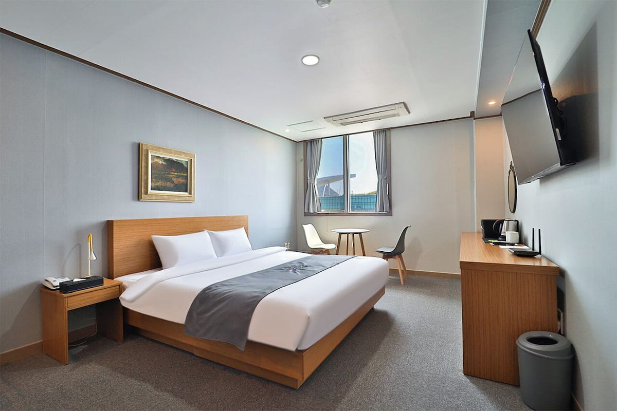 Habitación de hotel con una cama grande, mesita de noche, lámpara, escritorio, silla, televisor y ventana con cortinas.