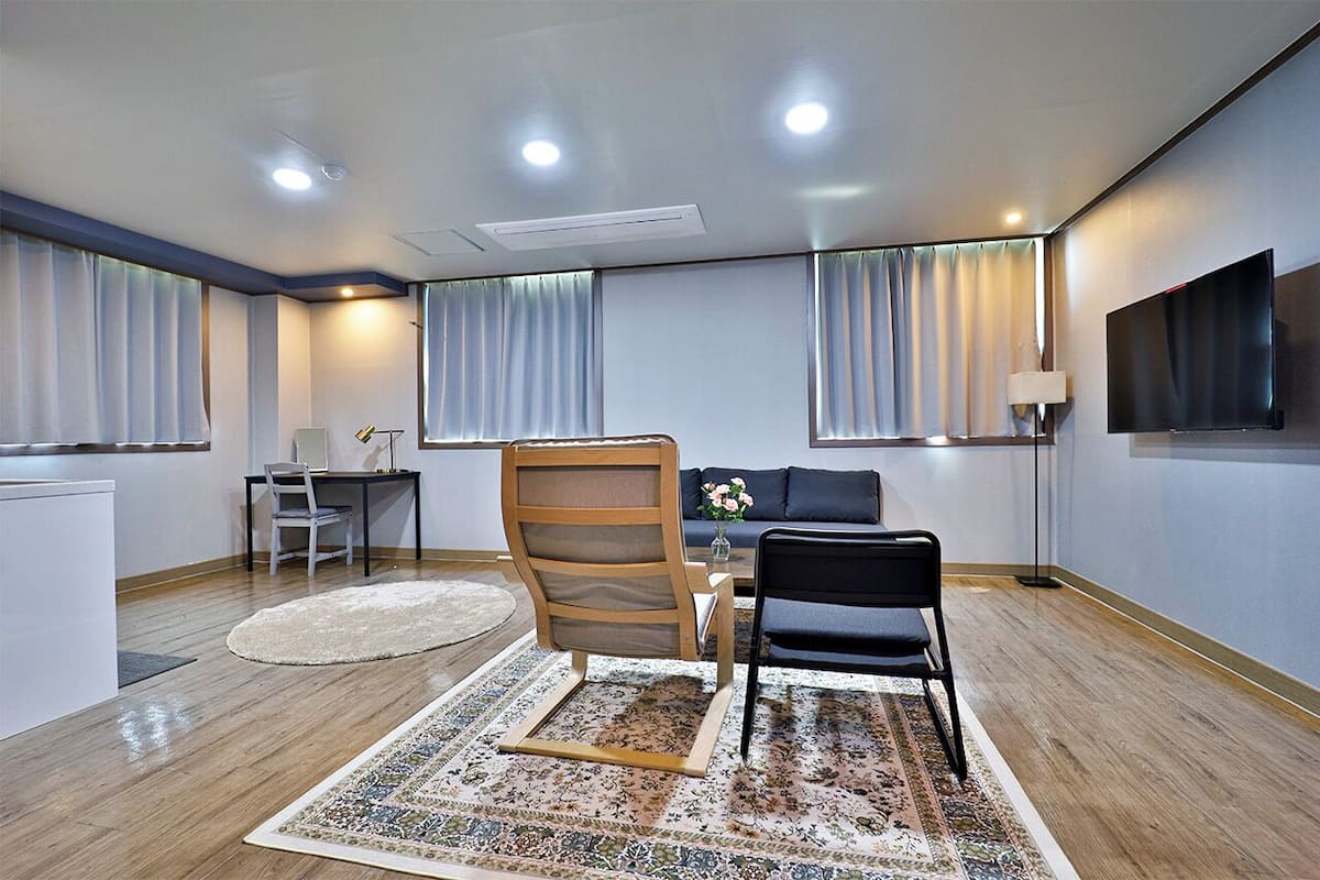 Habitación de hotel moderna con suelo de madera, un sofá, un escritorio con silla, televisión y un ventanal con cortinas.
