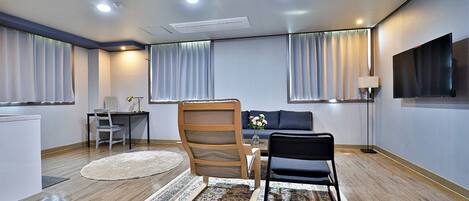 Habitación de hotel moderna con suelo de madera, un sofá, un escritorio con silla, televisión y un ventanal con cortinas.