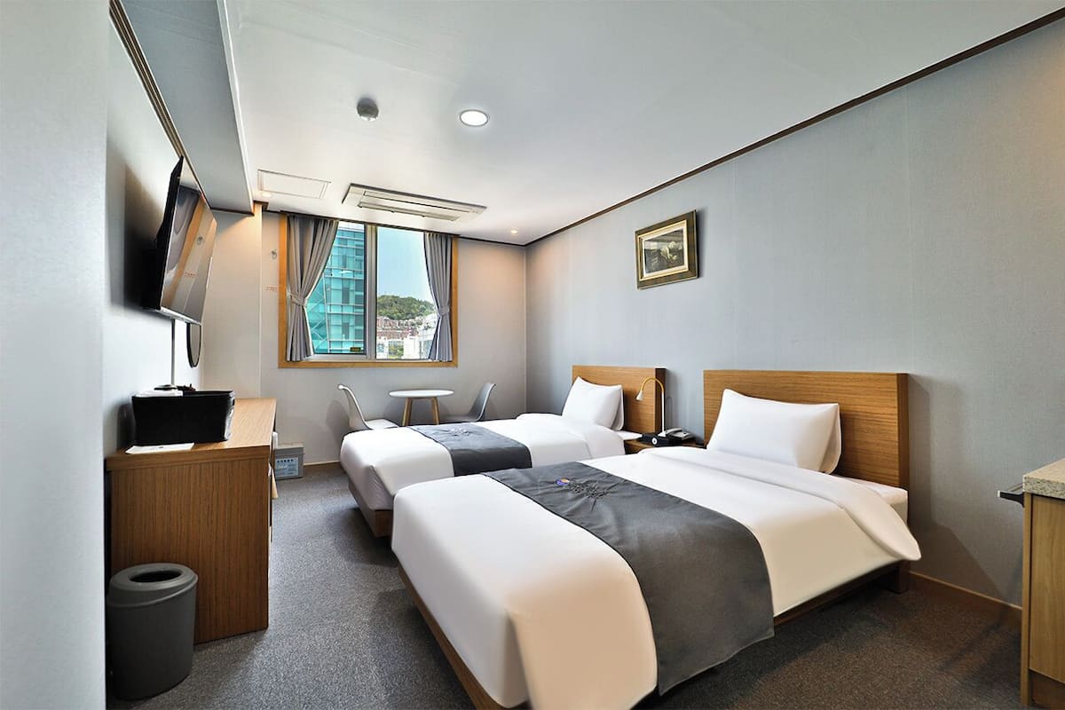 Habitación de hotel con dos camas, un escritorio, una silla, una mesita, televisor y ventana con vista a la ciudad.