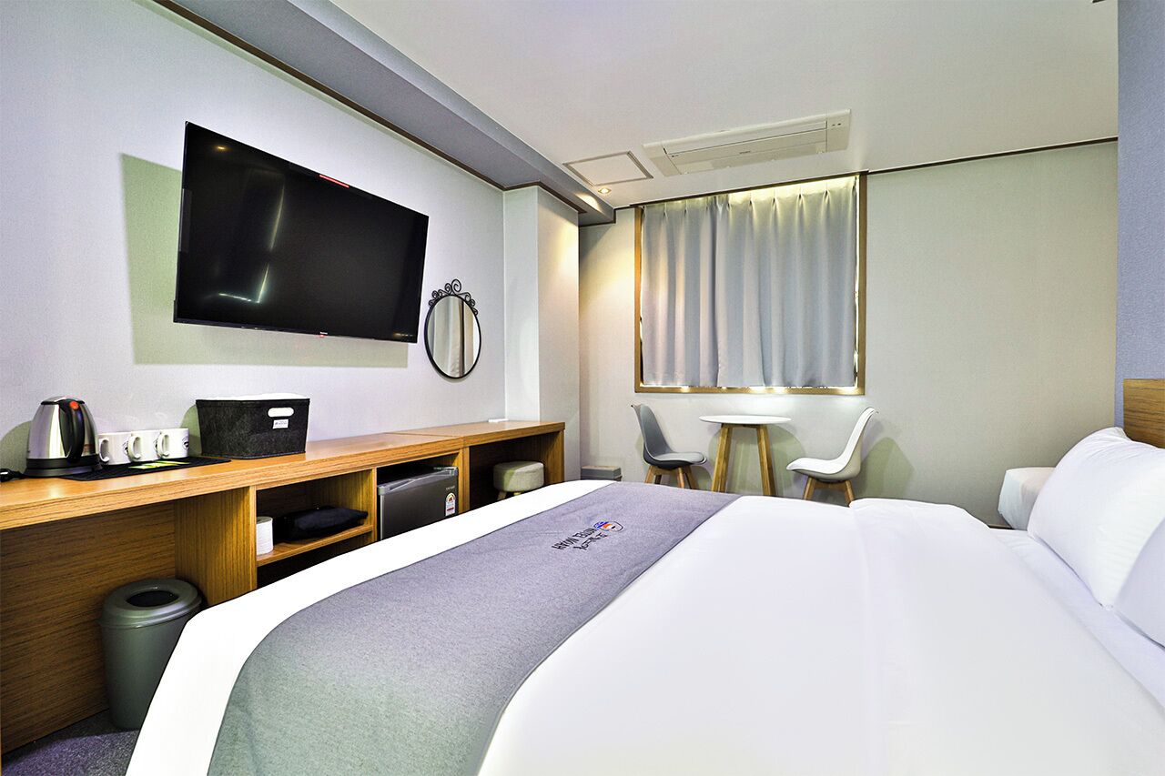A hotel room with a bed, a desk with a flat-screen TV, a small table with chairs, and a trash can.