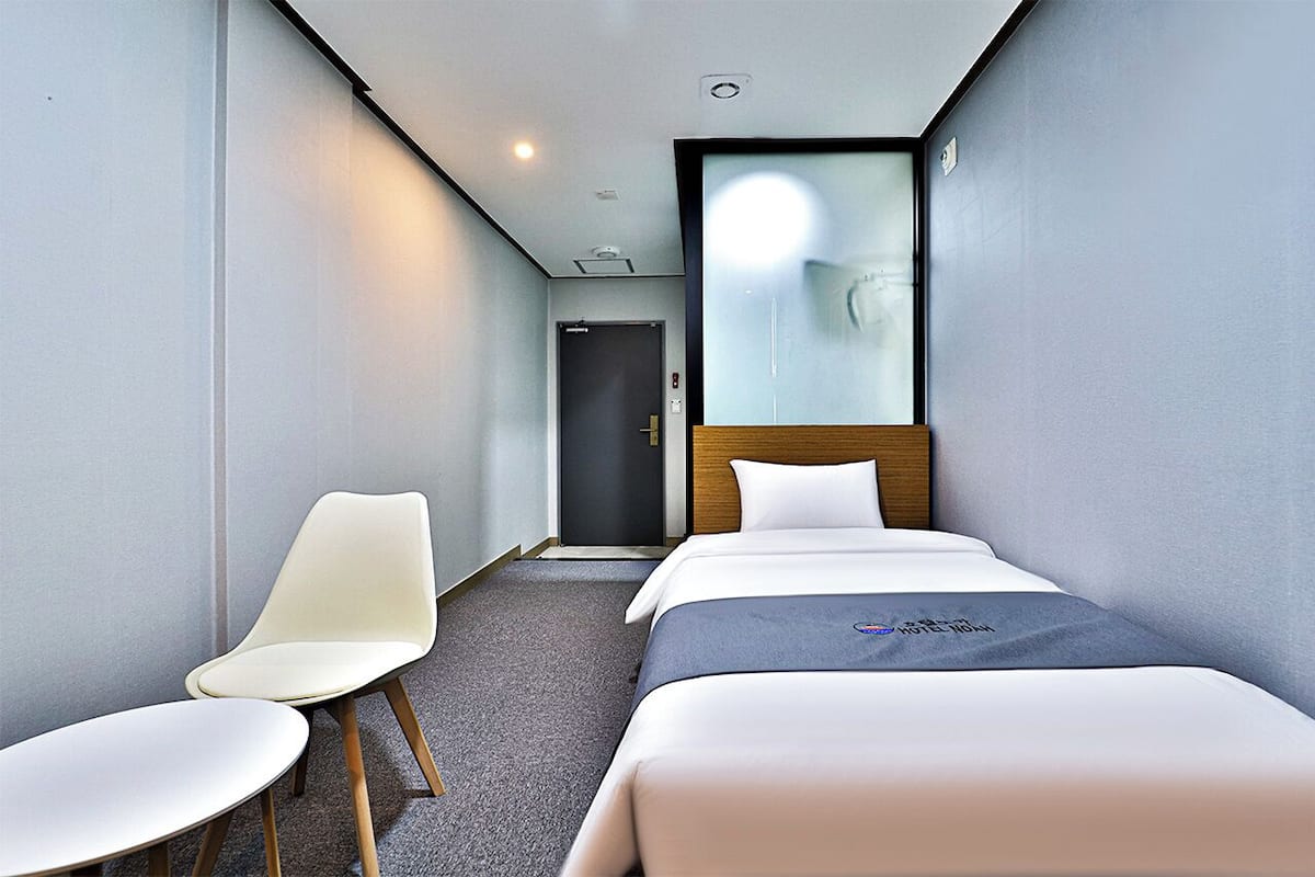 Habitación de hotel con dos camas individuales, una silla blanca, cabecera de madera y una puerta que da a otra habitación.