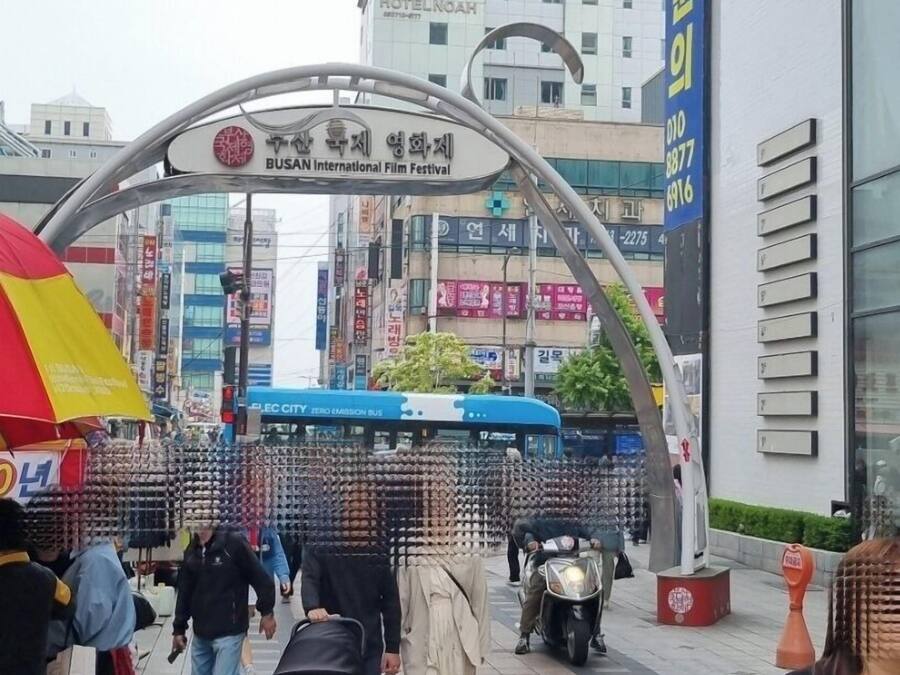 Zona peatonal con un cartel del Festival Internacional de Cine de Busan, un edificio moderno con un letrero azul y un arco de metal.
