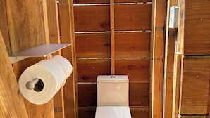 Un baño pequeño de madera con un inodoro, un rollo de papel higiénico, un cesto de basura negro y un desatascador.