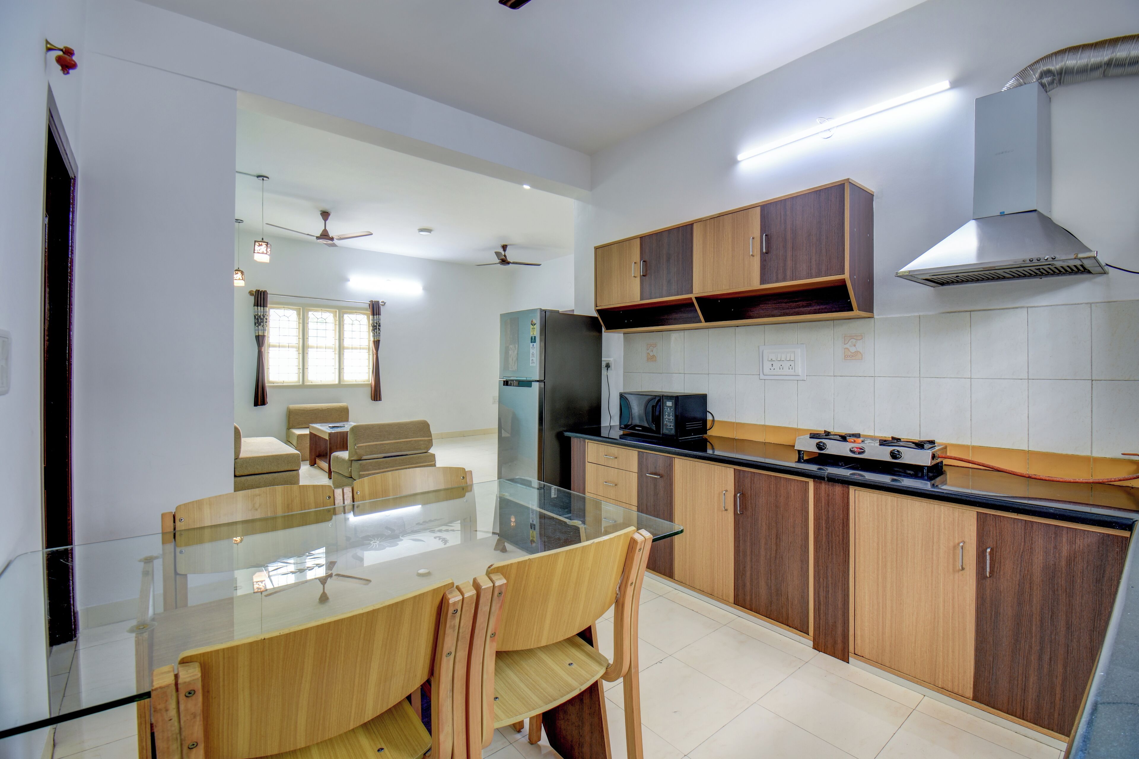 A modern kitchen with a glass dining table, wooden chairs, a refrigerator, microwave, and a gas stove.