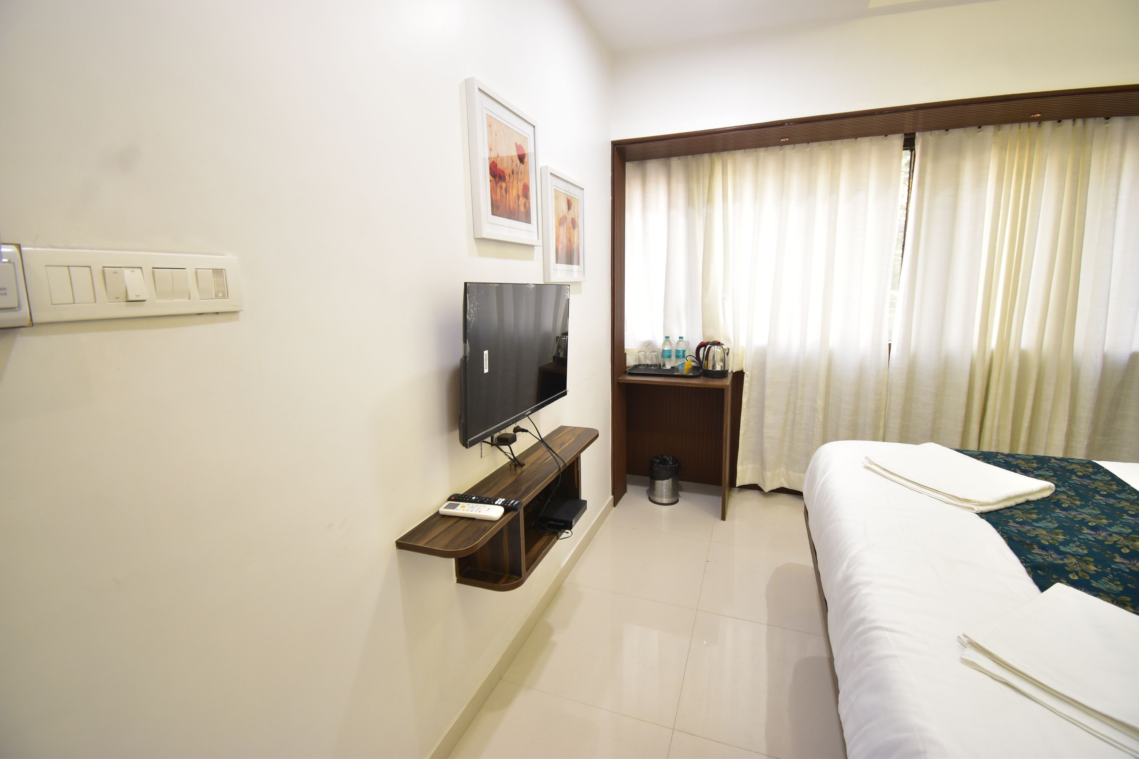 A hotel room with a bed, a TV mounted on the wall, a small table with a trash can, and a window with curtains.