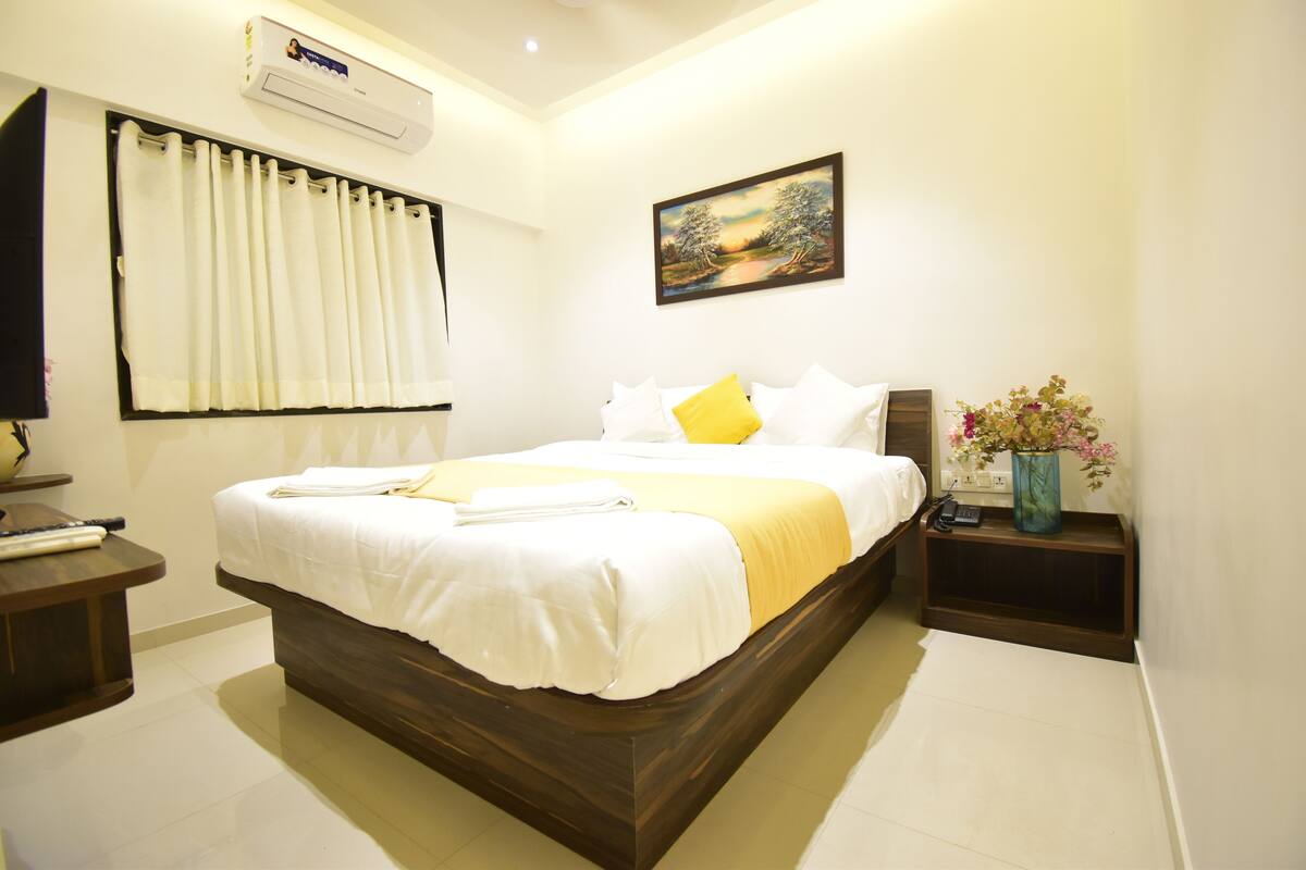 A neatly arranged hotel room with a large bed, a bedside table, a nightstand, a painting on the wall, and an air conditioning unit.