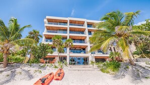 A multi-story hotel with a beachside location, featuring a central entrance with steps, surrounded by palm trees and two kayaks on the sand. - Tulum Beachfront Penthouse – Rooftop & Pool (Tulum)