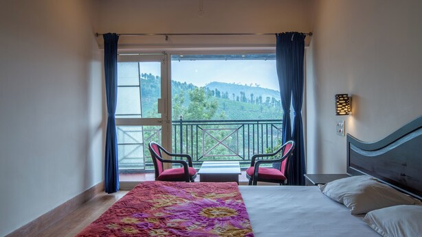 A bedroom with a bed, a window with curtains, a view of the outdoors, and a small table with chairs. - Misty Creek Homestay (Valparai)
