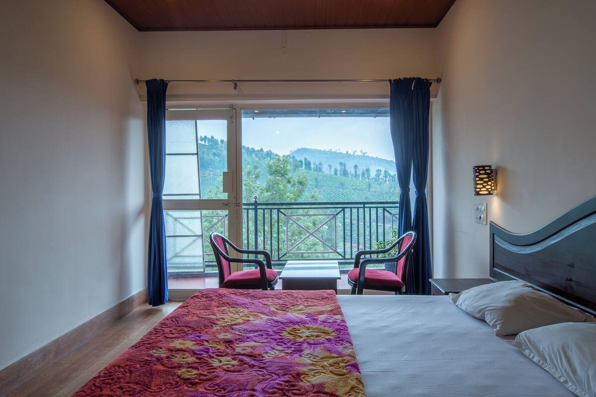 A bedroom with a bed, a window with curtains, a view of the outdoors, and a small table with chairs.