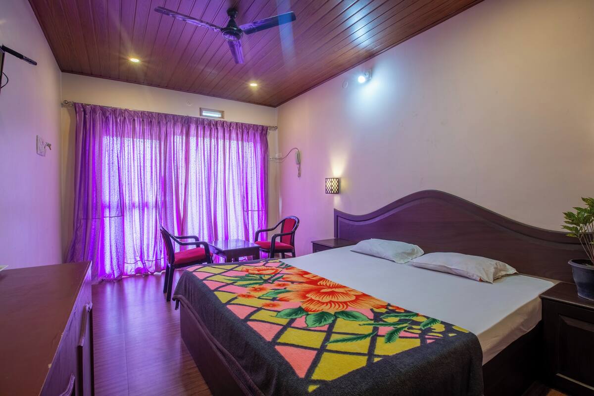 A hotel room with a bed, a desk, a chair, a ceiling fan, and a window with purple curtains.