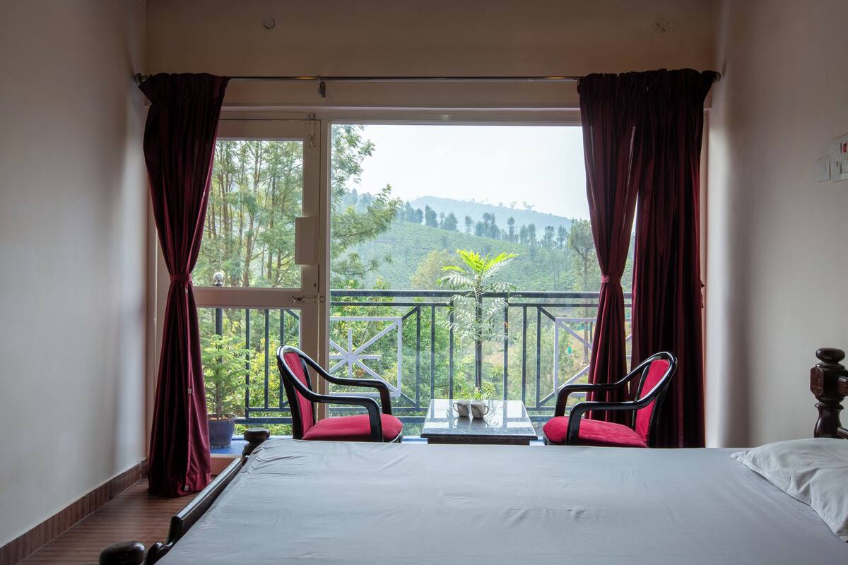 A bedroom with a large bed, two red chairs, a small table, and a view of a hilly landscape through a balcony with red curtains.