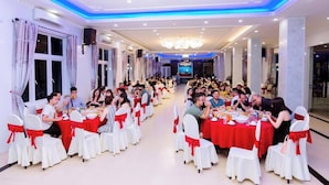 Un salón de banquetes con filas de mesas, sillas blancas con cintas rojas y una araña de cristal.