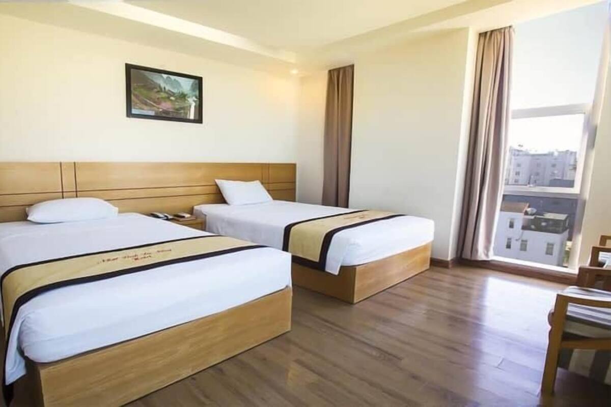 A hotel room with two beds, wooden headboards, a large window with curtains, and a framed picture on the wall.