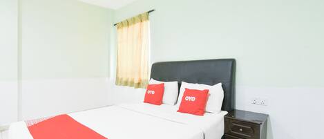 Habitación de hotel con cama, dos almohadas rojas, una mesita de noche y una ventana con cortinas.