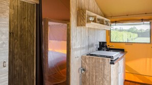 A room with a wooden door, a bed with a mosquito net, a kitchen area with a sink and countertop, and a large window with a view of a vineyard. - Cozy Glamping in Mierlo (Mierlo)