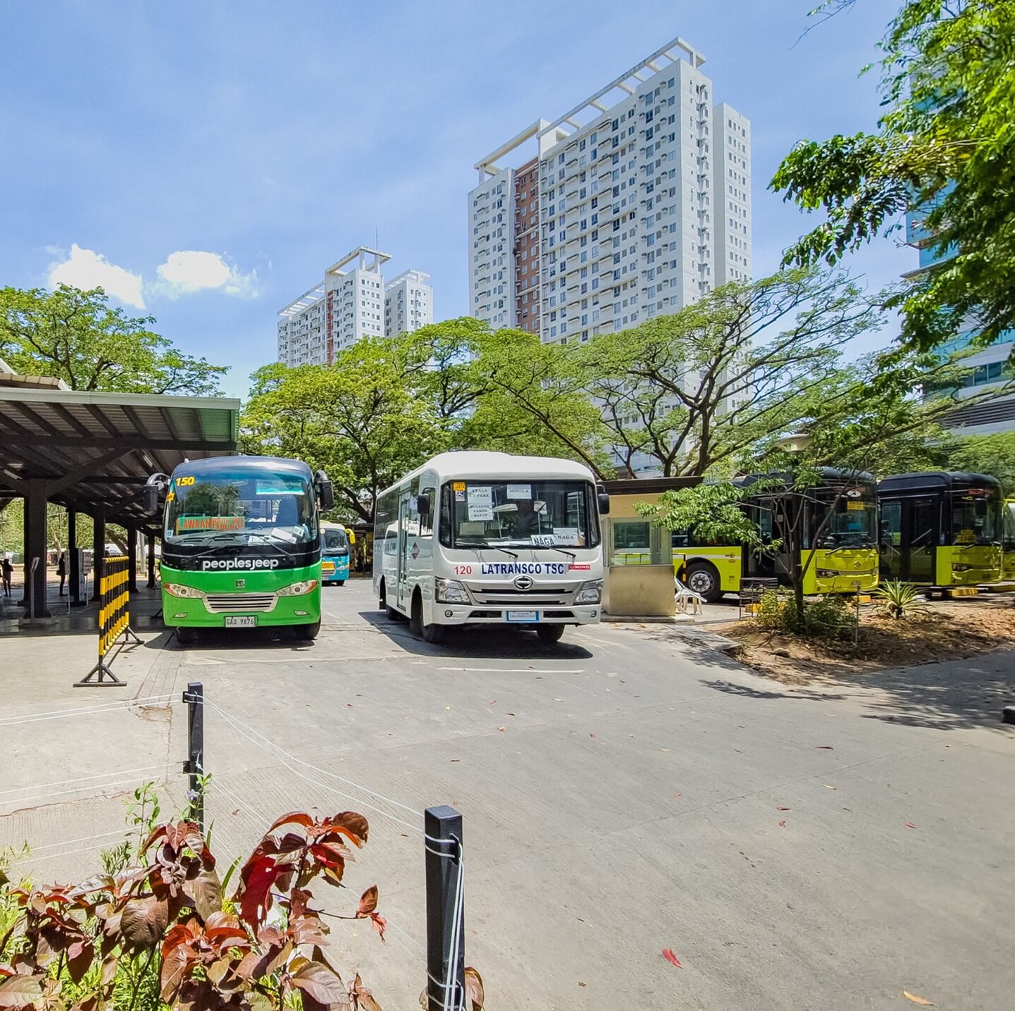 Una estación de autobuses con dos vehículos, un área de espera techada y edificios residenciales altos al fondo.