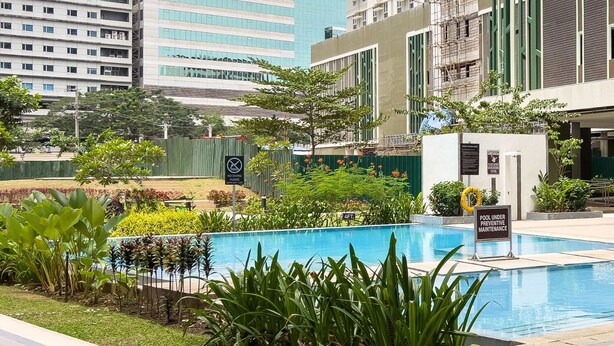 A modern hotel with a swimming pool surrounded by lush greenery and urban buildings in the background.