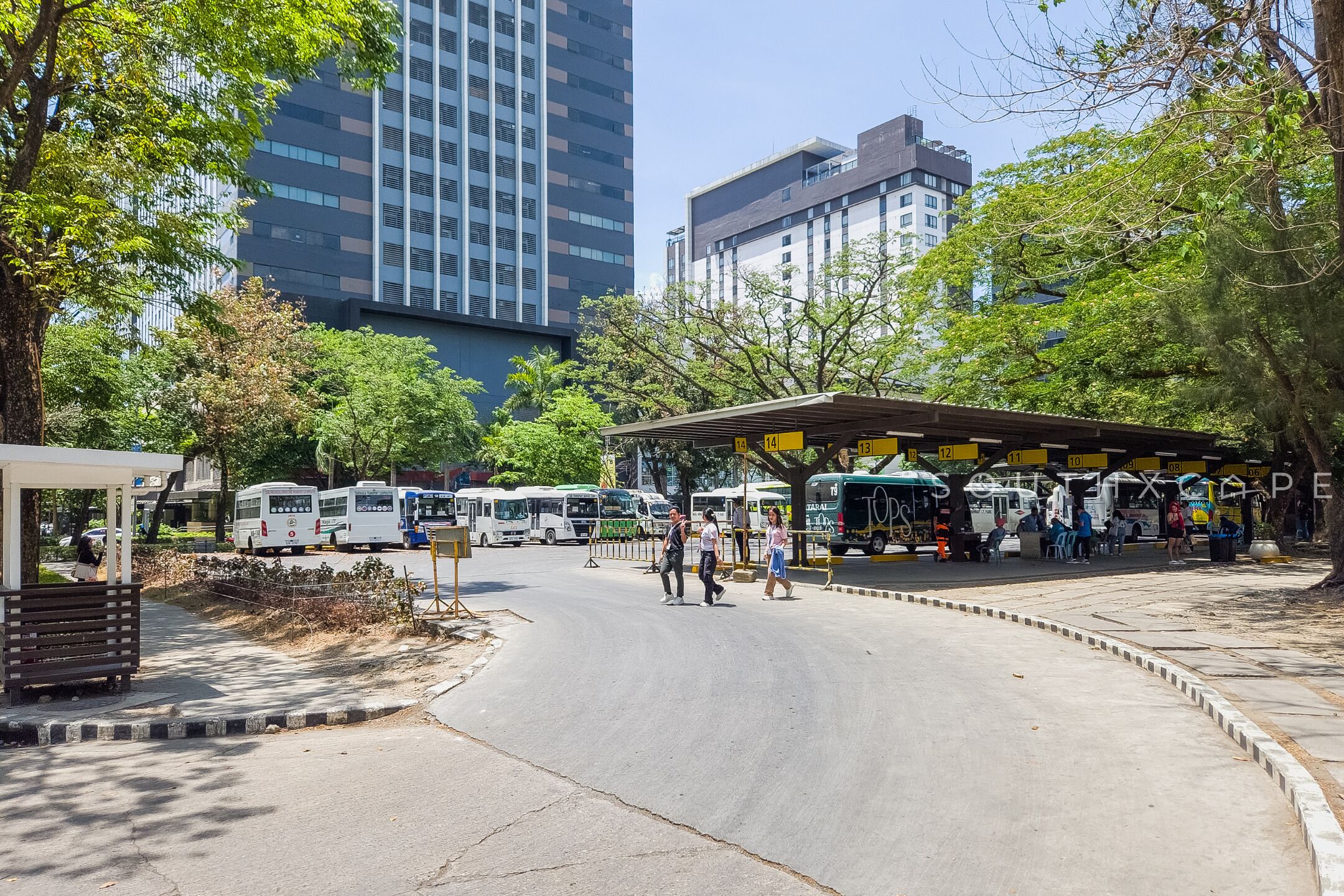 Una estación de autobuses con varios vehículos, una zona de espera techada y edificios altos al fondo.