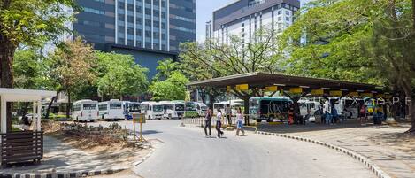 Una estación de autobuses con varios vehículos, una zona de espera techada y edificios altos al fondo.
