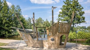 A wooden pirate ship play structure with a ladder, a steering wheel, and a flag.