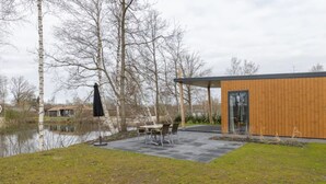 A modern building with a wooden exterior and a glass door, situated next to a body of water and a grassy area with a table and chairs.