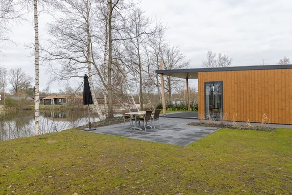 Un edificio moderno con fachada de madera y puerta de vidrio, ubicado junto a una masa de agua y un área verde con una mesa y sillas.