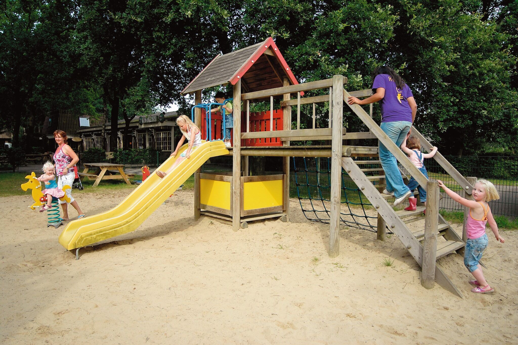 A playground with a yellow slide, wooden climbing structures, and a small house-like playhouse.