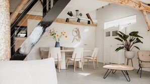 A modern loft-style living room with a dining area, featuring exposed wooden beams, a white sofa, a round coffee table, and a large potted plant.