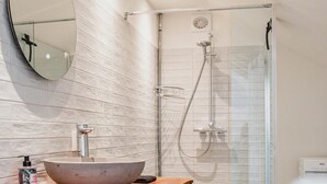 A modern bathroom with a large oval mirror, a wooden vanity with a bowl sink, a shower with a glass door, and a washing machine.