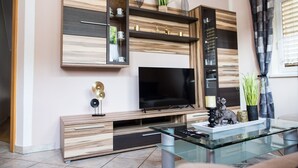 A modern living room with a flat-screen TV, wooden shelving, and a glass coffee table. - Cosy apartment in Arnoldstein (Arnoldstein)