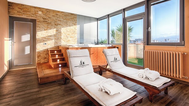 A modern spa room with wooden flooring, a stone wall, and large windows offering a view. There are two sunbeds with towels and a wooden staircase.