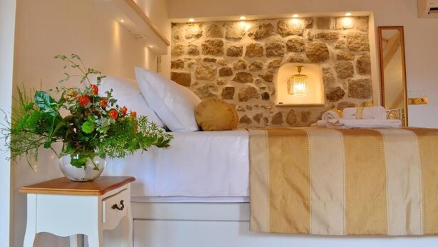 A bedroom with a stone wall, a bed with white bedding, a bedside table with a vase of flowers, and a mirror on the wall. - Villa madonna della Strada A5 (Dubrovnik)