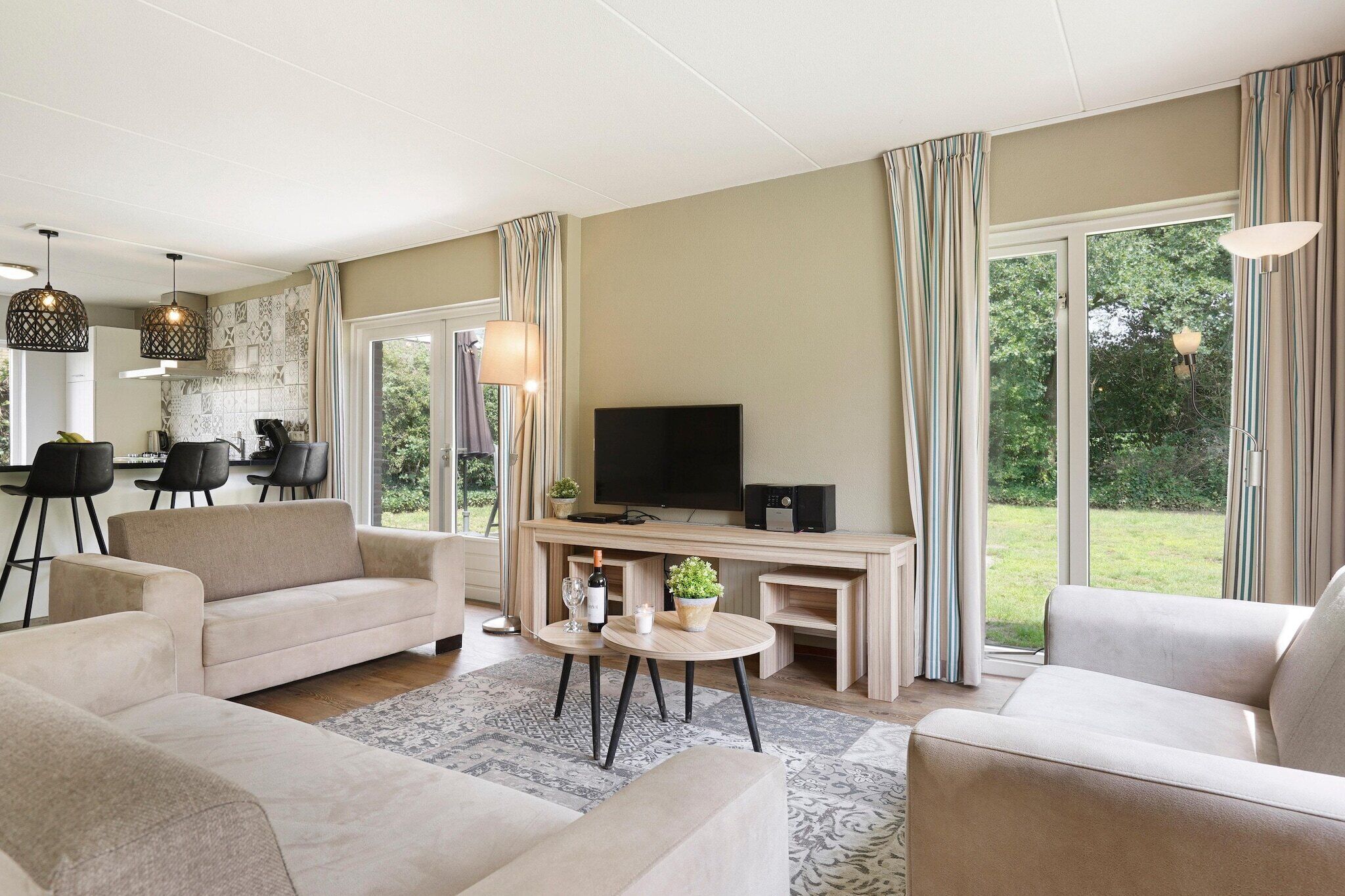 Een moderne woonkamer met een bank, salontafel en televisie. Er is een eethoek met stoelen en een tafel. Grote ramen bieden uitzicht op de omgeving.