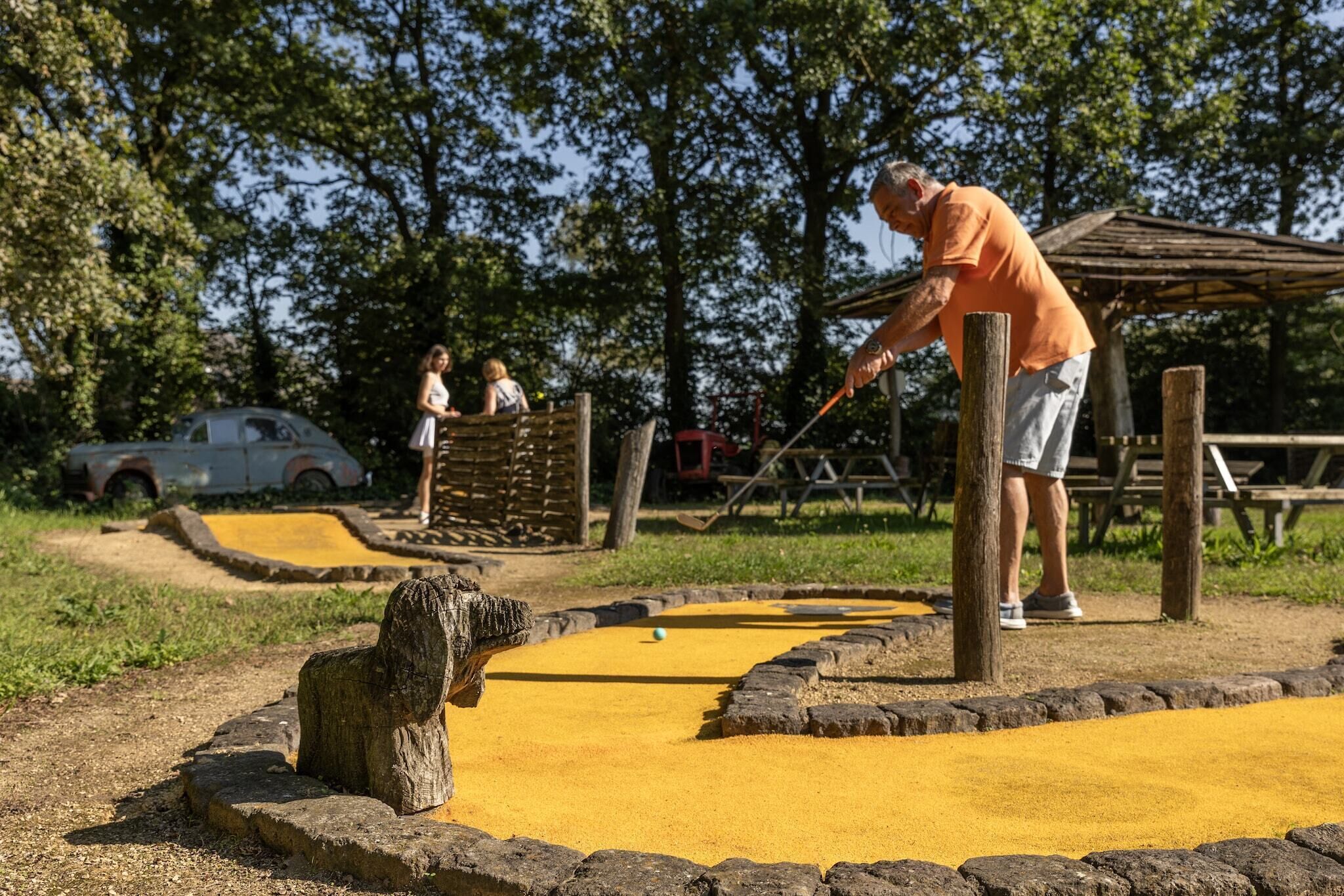 A man playing shuffleboard in an outdoor setting with a wooden structure and a yellow playing surface.