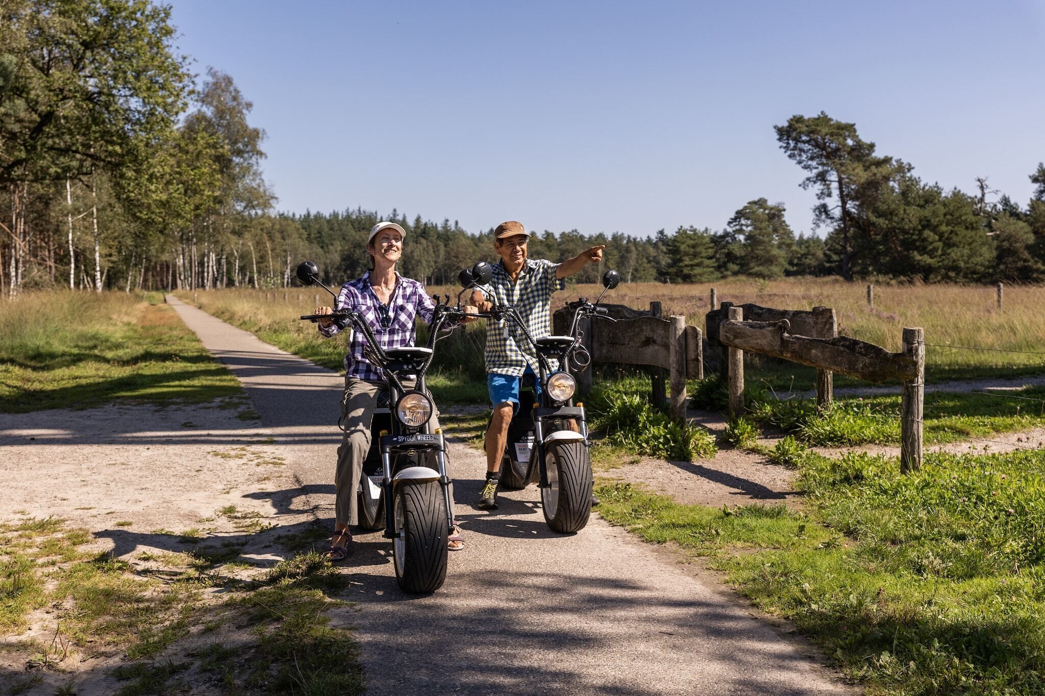 Deux personnes sur des motos sur une route de campagne, avec une clôture en bois et des arbres en arrière-plan.