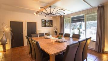 Une salle à manger avec une table en bois, des chaises noires, un lustre et une vue sur l’extérieur grâce à de grandes fenêtres.