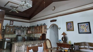 Un vaste espace de vie doté d’un haut plafond, de poutres en bois et d’un ventilateur de plafond. On y trouve un coin cuisine avec des placards, ainsi qu’un coin repas avec des chaises et une table.