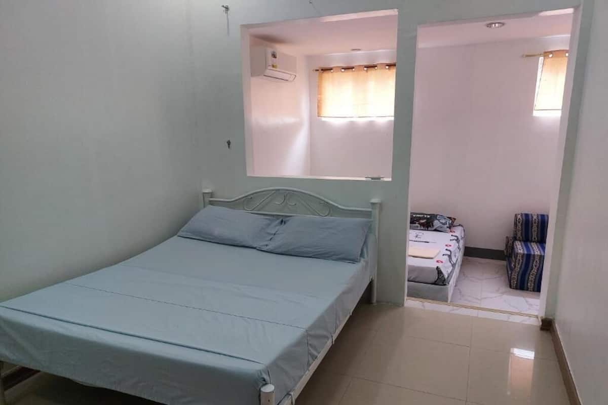 Une chambre avec un lit simple, un cadre en métal, une fenêtre avec des rideaux, une petite table et une chaise.