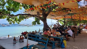 Un espace de restauration en plein air, avec des tables et des chaises disposées sous un arbre, offrant une vue sur la mer.