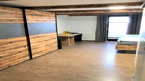 Une pièce avec un plancher en bois, un lit, une table avec un vase et une fenêtre avec des rideaux.