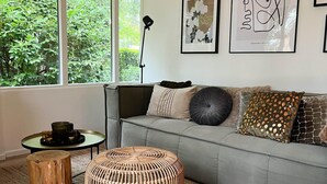 Een moderne woonkamer met een grijze bank, een rond geweven pouf, een klein rond tafeltje met kaarsen, en een vloerlamp.