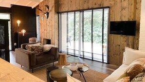 Un salón moderno con una pared de madera, un ventanal con persianas, un sofá, una mesa de centro y un televisor montado en la pared.