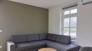 Ein modernes Wohnzimmer mit einem grauen Ecksofa, zwei kleinen runden Tischen und einem großen Fenster mit Blick ins Freie.