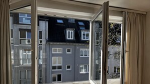Une pièce dotée d’une grande fenêtre, d’un radiateur et offrant une vue sur un immeuble à plusieurs étages avec des balcons.